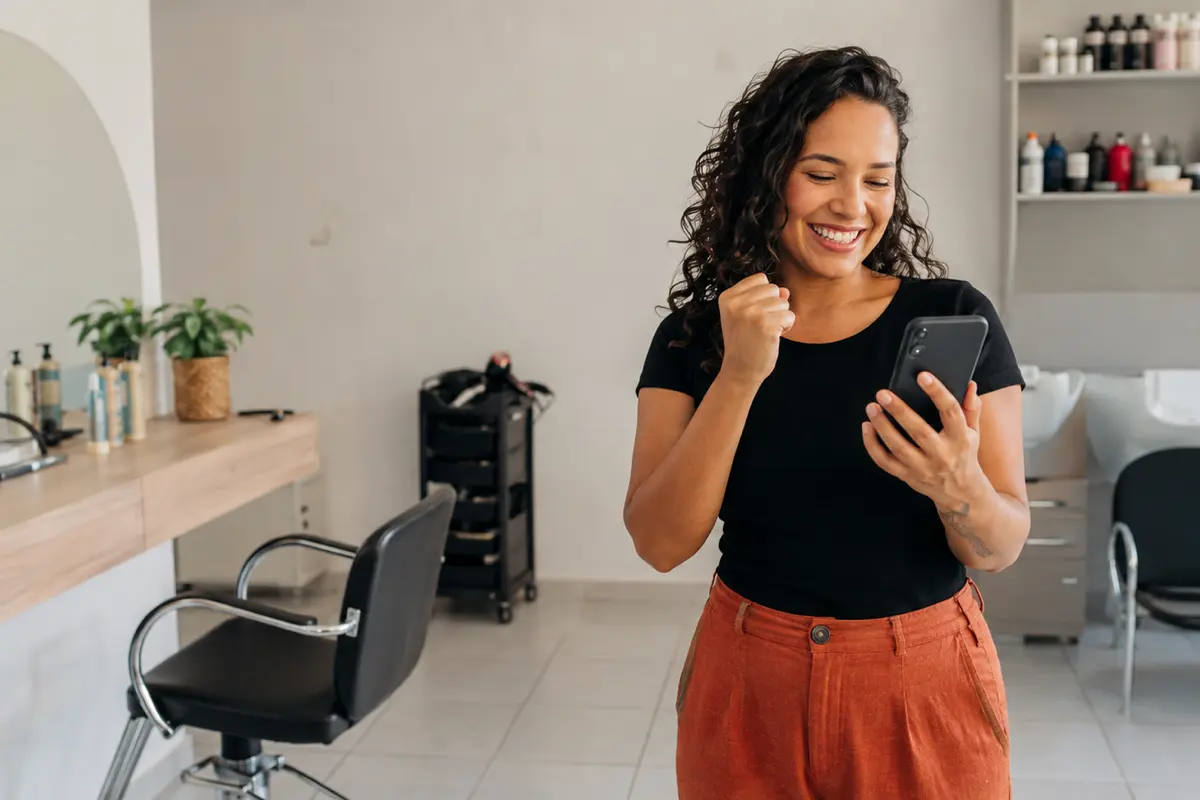 Dona de salão recém-aberto sorri com entusiasmo enquanto organiza a divulgação no celular; ilustração gerada por IA