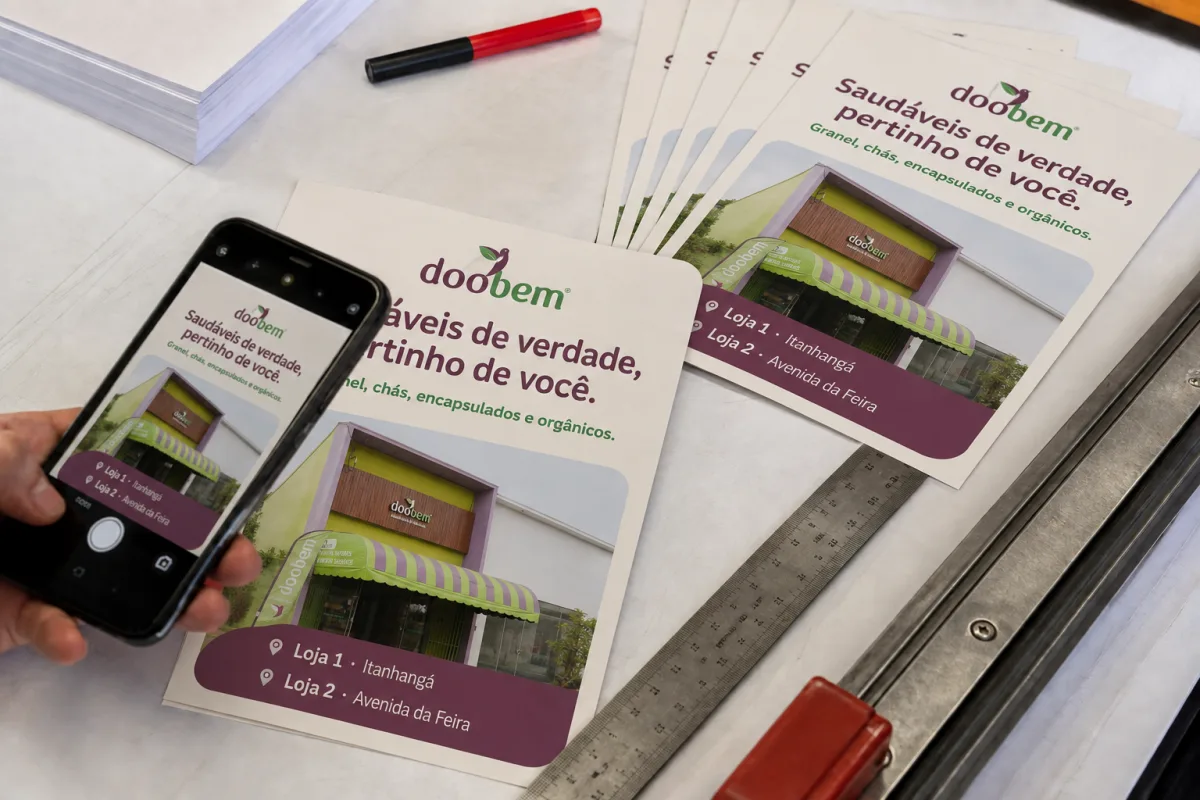 Panfletos da Doobem com a fachada das lojas sendo conferidos pelo celular em mesa de gráfica; ilustração gerada por IA com marca oficial